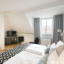 Ein helles, modernes Hotelzimmer mit einem Doppelbett, einer geometrisch gemusterten Decke, einem Fernseher auf einem Ständer, zwei Stühlen, einem kleinen Tisch, einem Schreibtisch und großen Fenstern mit grauen Vorhängen, die das Tageslicht hereinlassen.