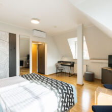 Ein helles, modernes Hotelzimmer mit zwei Einzelbetten, einer Wandkarte, einem Schreibtisch am Fenster, einem Fernseher auf einem Schrank, einem Kleiderschrank mit Spiegel und Holzfußboden. Der Raum ist von natürlichem Licht durchflutet.