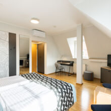 Ein helles, modernes Hotelzimmer mit zwei Einzelbetten, einer Wandkarte, einem Schreibtisch am Fenster, einem Fernseher auf einem Schrank, einem Kleiderschrank mit Spiegel und Holzfußboden. Der Raum ist von natürlichem Licht durchflutet.