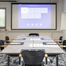 Ein moderner Konferenzraum mit einem großen Tisch, acht Stühlen, Notizblöcken, einem Flipchart und einem Projektionsbildschirm, auf dem eine Präsentation gezeigt wird. Ein Fenster mit Jalousien und ein gemusterter Teppich sind zu sehen. Der Raum ist leer.