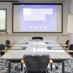 Tagungsraum 3 Ein moderner Konferenzraum mit einem großen Tisch, acht Stühlen, Notizblöcken, einem Flipchart und einem Projektionsbildschirm, auf dem eine Präsentation gezeigt wird. Ein Fenster mit Jalousien und ein gemusterter Teppich sind zu sehen. Der Raum ist leer.